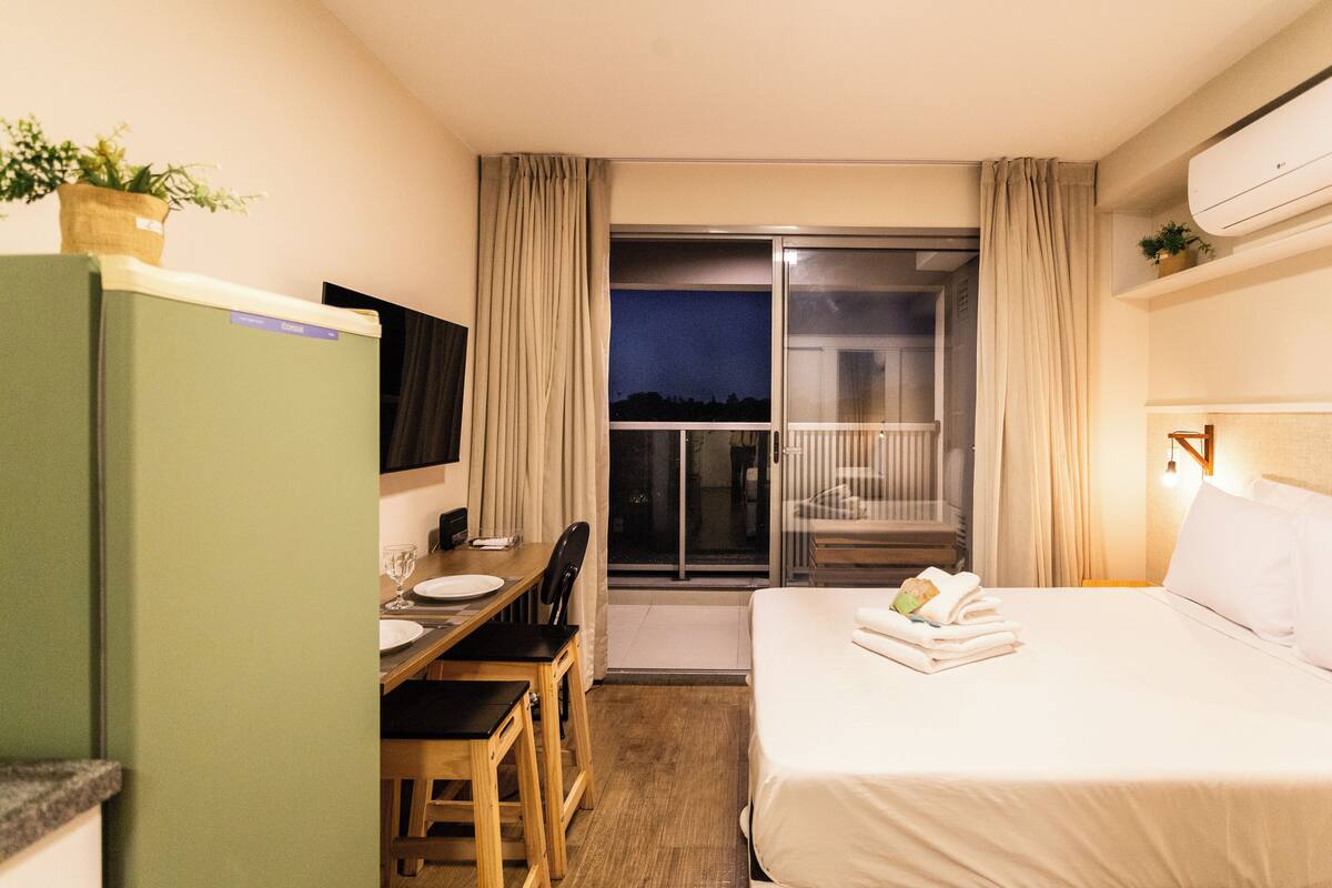 Habitación de hotel con cama, escritorio, silla, televisor y balcón con cortinas.