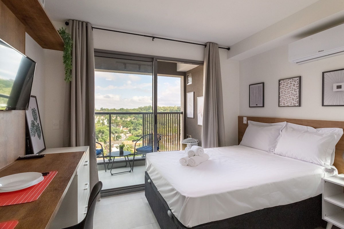 Habitación de hotel con cama, escritorio con televisor, balcón con una silla y vista a la ciudad.