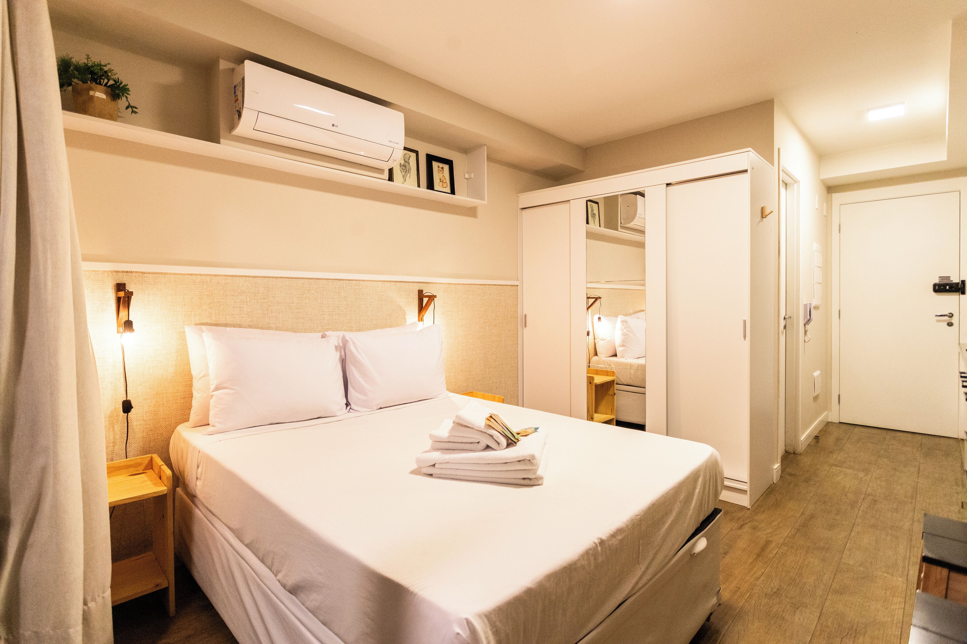 A hotel room with a neatly made bed, a bedside table with a lamp, a wardrobe, and a small shelf with a plant and framed pictures.