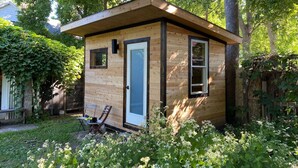 Una pequeña cabaña de madera con puerta y ventanas, rodeada de follaje y árboles.