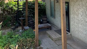 Una terraza de madera con una escalera que conduce a un balcón en un segundo piso, rodeada de follaje y un edificio con pared texturizada.