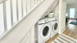 Un espace buanderie comprenant deux machines à laver à chargement frontal, un panier à linge et un escalier avec des rampes blanches.