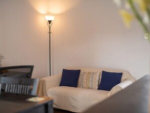 Un salon avec un canapé beige, deux coussins bleus, un plaid à motifs et une lampe sur pied.
