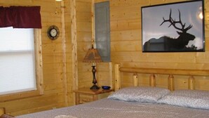 Ein Zimmer einer Holzblockhütte mit einem Bett, einem Deckenventilator, einer Uhr, einer Lampe und einer Hirsch-Silhouetten-Grafik.
