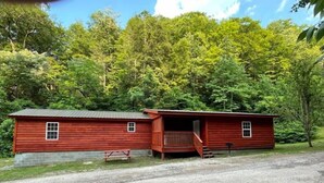 A red wooden cabin with a porch and a gravel driveway, surrounded by dense forest. - 3BR, 2 Bath Cabin with full kitchen, living room, and hot tub. (Putney)