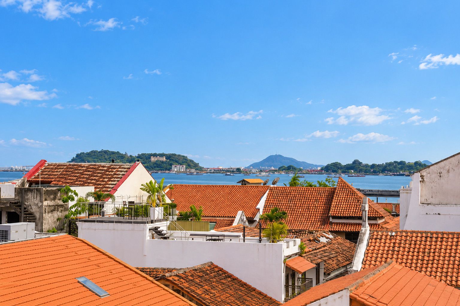 Un paisaje urbano costero con edificios de techos de teja rojiza, un cielo azul despejado y una montaña distante.