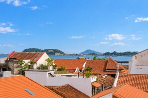 Paisagem costeira urbana com edifícios de telhados de terracota, céu azul claro e montanha distante.