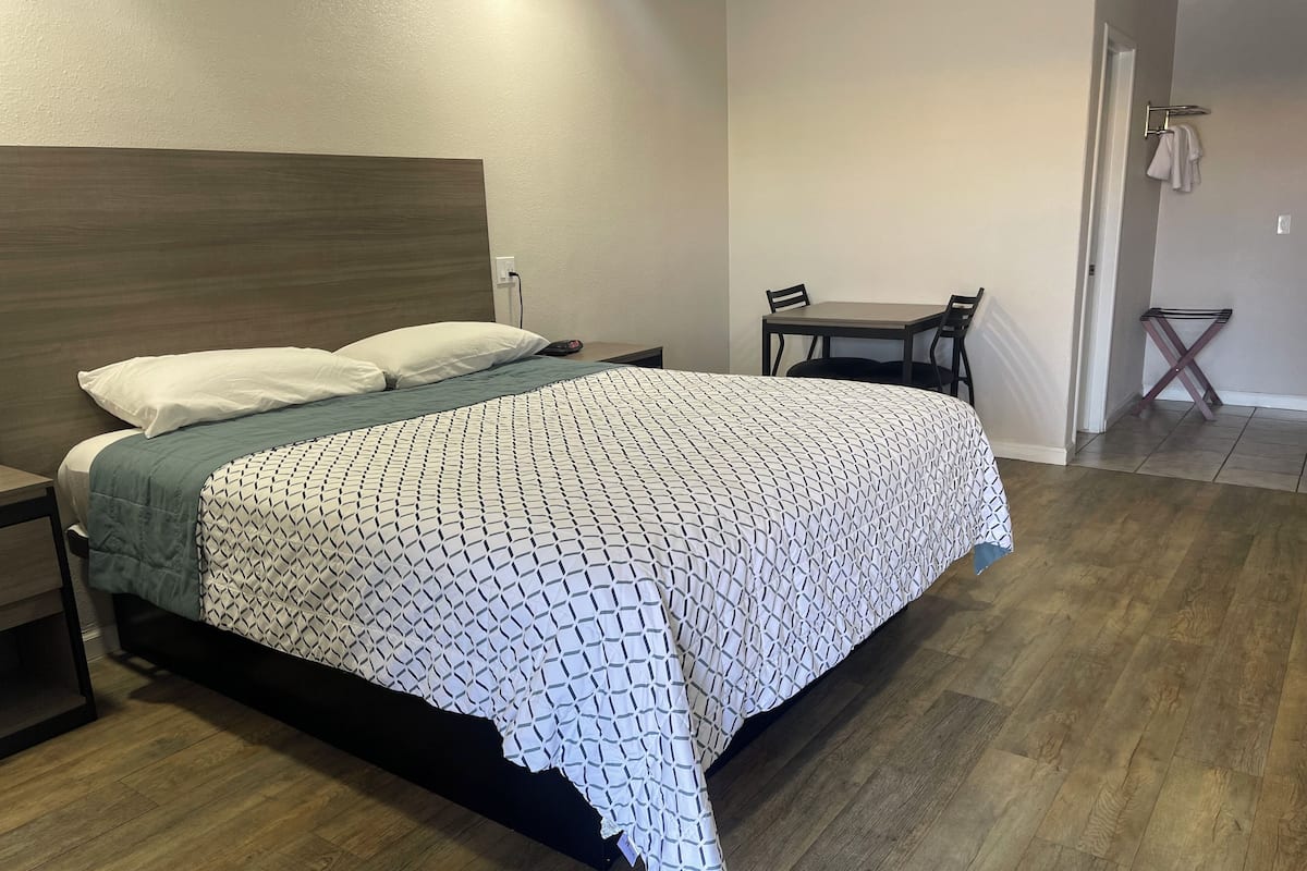 A neatly made bed with a patterned bedspread, a bedside table, and a chair in a room with wooden flooring and a plain wall.