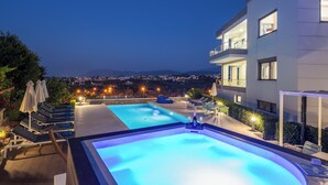 Un área de piscina moderna con sillones de descanso y sombrillas, rodeada de un jardín ajardinado y vistas a un paisaje urbano nocturno.