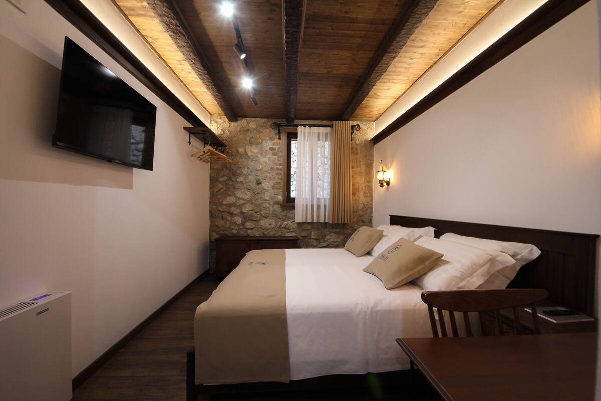 A bedroom with a bed, a TV mounted on the wall, a stone wall, and a wooden ceiling.