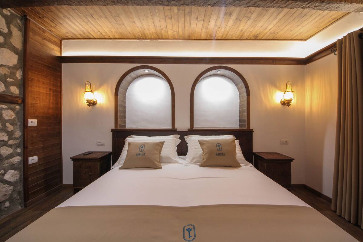 A hotel room with a large bed, two nightstands, a stone wall, wooden ceiling, and arched windows.