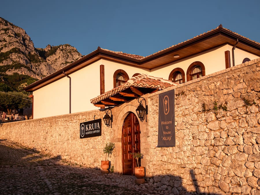 Edifici de parets de pedra amb teulada de rajola, una porta de fusta i dues plantes en testos. Un cartell diu "KRUA ALBERGO DI FUSO" i un altre diu "Benvinguts a House 'KALAJI'".