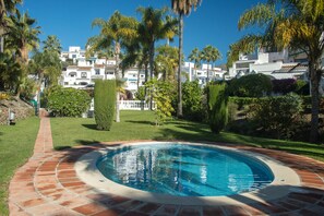 A swimming pool with a brick pathway, surrounded by well-maintained grass and palm trees, with a building in the background. - Tetuan 31 - El Oasis de Capistrano! (Nerja)