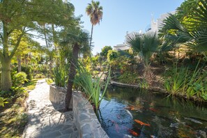 Un jardin avec un sentier en pierre, un bassin avec des carpes koï et diverses plantes tropicales.