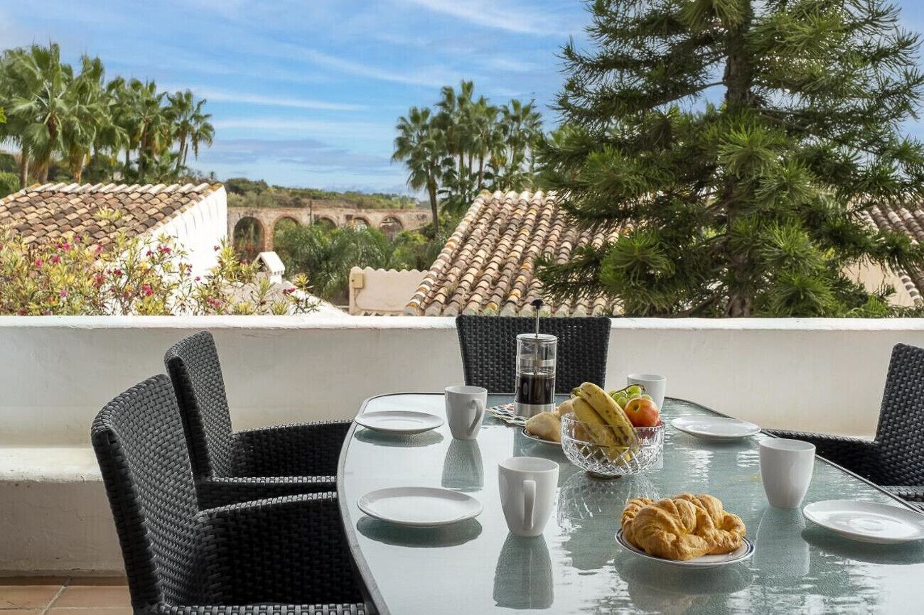 Een eettafel met stoelen, een croissant, fruit en een glas water op een balkon met uitzicht op palmbomen en een brug.