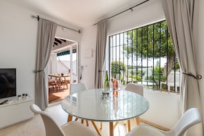 A modern room with a glass table, white chairs, a TV, and a balcony with a view.