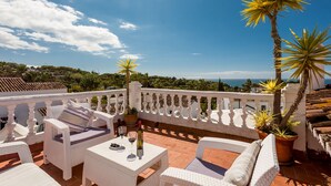 Eine Terrasse mit weißen Korbmöbeln, einem Tisch mit einer Flasche und Gläsern sowie Topfpflanzen.