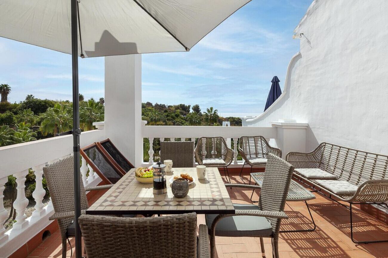 Eine Terrasse im Freien mit Korbmöbeln, einem gefliesten Tisch und einem weißen Sonnenschirm.