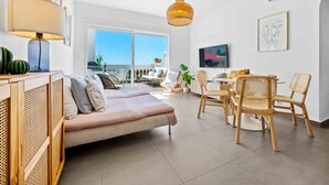 En moderne stue med sofa, spisebord, fletstole, fjernsyn og et stort vindue.