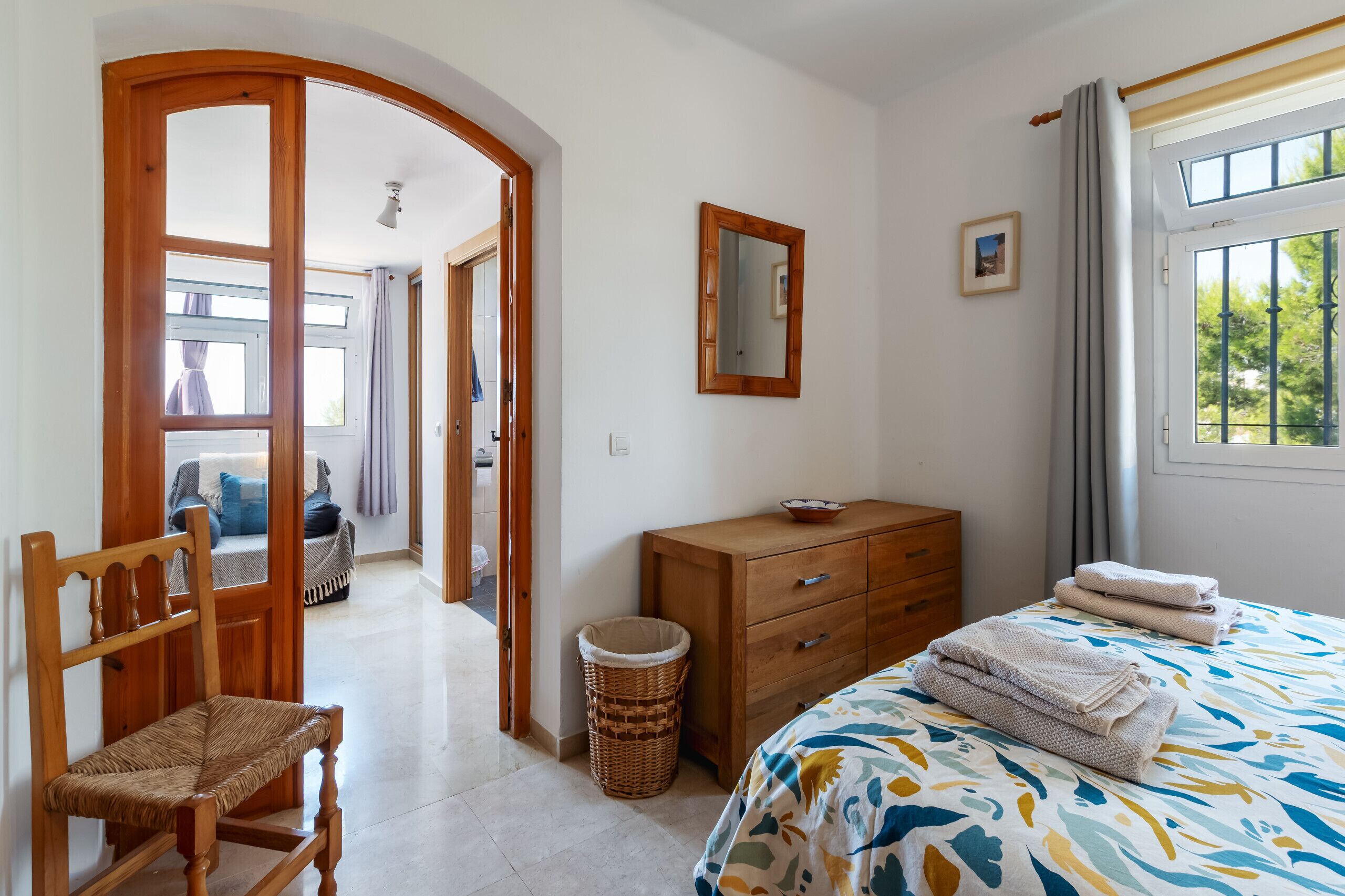 Une chambre à coucher avec un lit, une commode en bois, une chaise, un miroir et une fenêtre avec des rideaux.