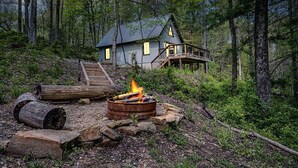 Une cabane dotée d’une terrasse et d’un foyer extérieur, située dans un cadre forestier.