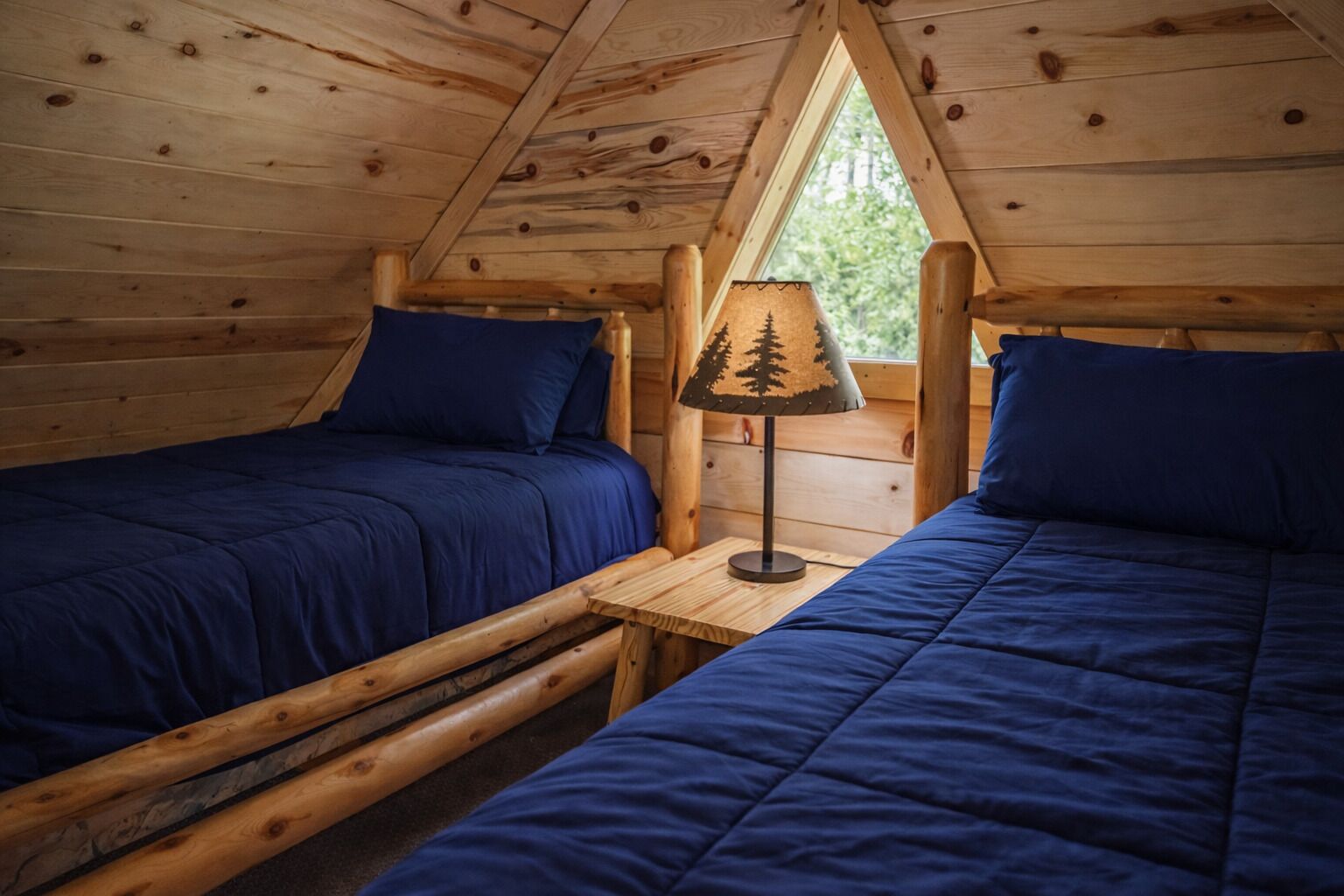 Une cabane avec deux lits en bois, une table de chevet avec une lampe et une fenêtre donnant sur la forêt.