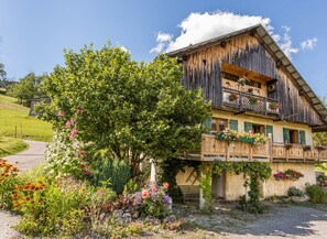 Situated in a rural setting, this chalet features a balcony with flower baskets and a well-maintained garden with a large tree.