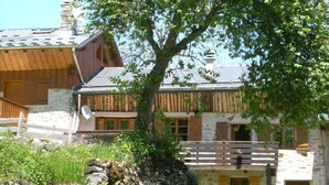Un chalet rustique avec une façade en pierre, des balcons en bois et un grand arbre au premier plan.