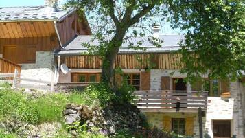 Un chalet rustique avec une façade en pierre, des balcons en bois et un grand arbre au premier plan.