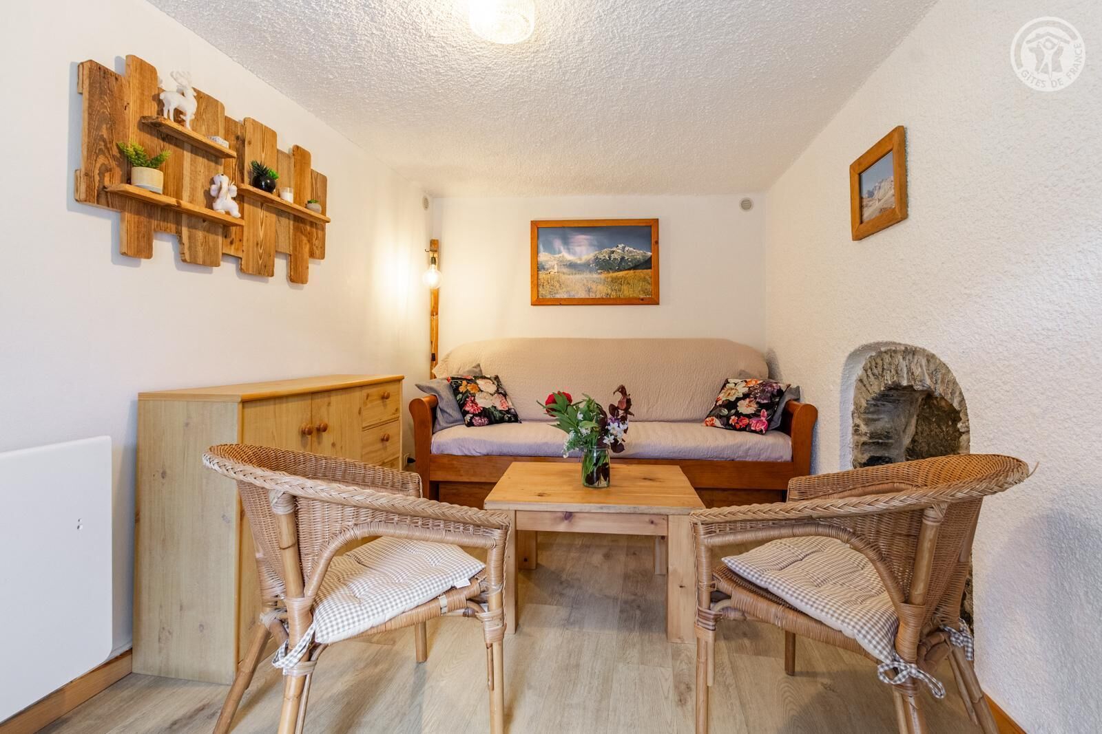 Quarto acolhedor com sofá, duas cadeiras de vime, mesa de madeira, lareira e prateleira fixada na parede com objetos decorativos.