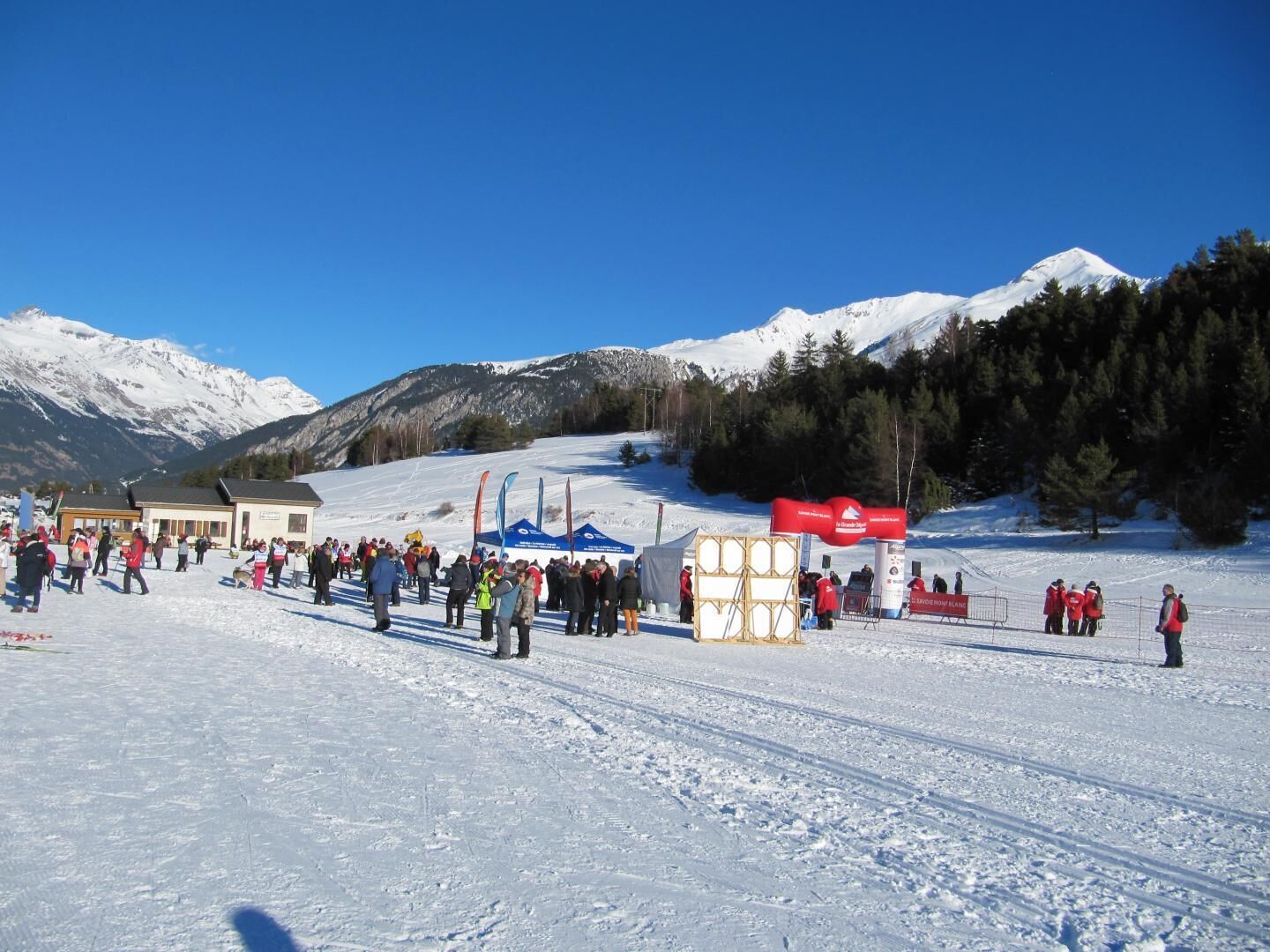 A snowy outdoor area with a crowd of people, tents, and a mountain backdrop.