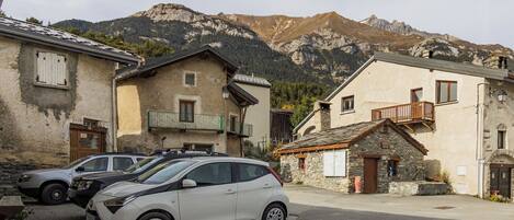 Uma fileira de edifícios de pedra tradicionais com varandas, carros estacionados e montanhas ao fundo.