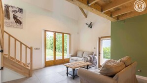 A spacious living room with a wooden staircase, a beige sofa, a coffee table, and a large sliding glass door leading to an outdoor area. - LA BERGERIE DE CLAUDE (LE BOURGET DU LAC)