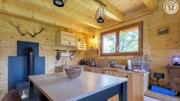 Une cabane en bois rustique dotée d’un coin cuisine comprenant une grande table en bois, un poêle et une fenêtre donnant sur la forêt.