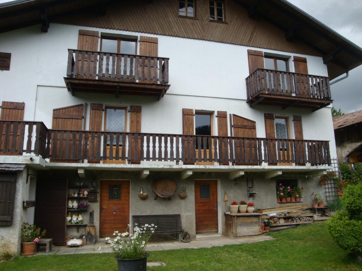 A two-story building with wooden balconies, a front door, and a small garden area with potted plants.