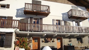 A multi-story building with wooden balconies, a snow-covered ground, and a small garden area with flowers. - LES GENTIANES - REGUET (NOTRE DAME DE BELLECOMBE)
