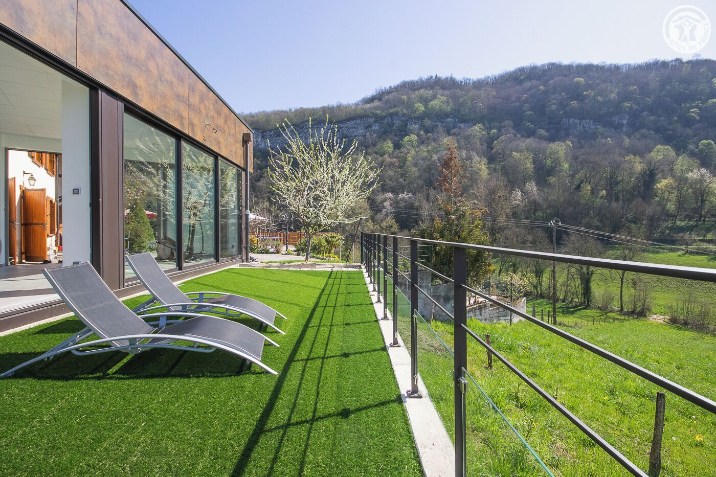 Una casa moderna con balcón que da a un césped verde exuberante y una ladera boscosa.