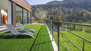 Ein modernes Haus mit Balkon, das auf eine üppige grüne Wiese und einen bewaldeten Hang blickt.