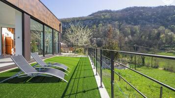 A modern house with a balcony overlooking a lush green lawn and a forested hillside.