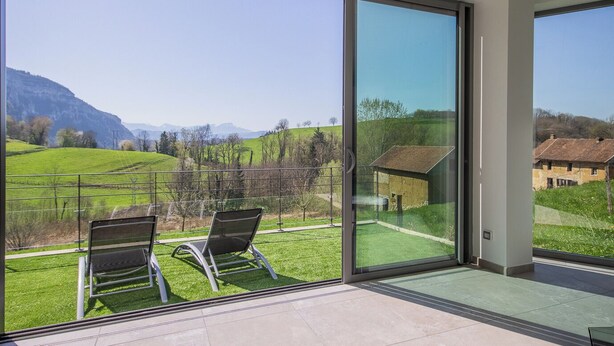 Ein modernes Zimmer mit großen Glasfenstern und Blick auf eine grüne Landschaft sowie Berge.
