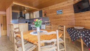 Un intérieur en bois comprenant une table à manger, des chaises, un vase avec des fleurs, un coin cuisine et un téléviseur fixé au mur.