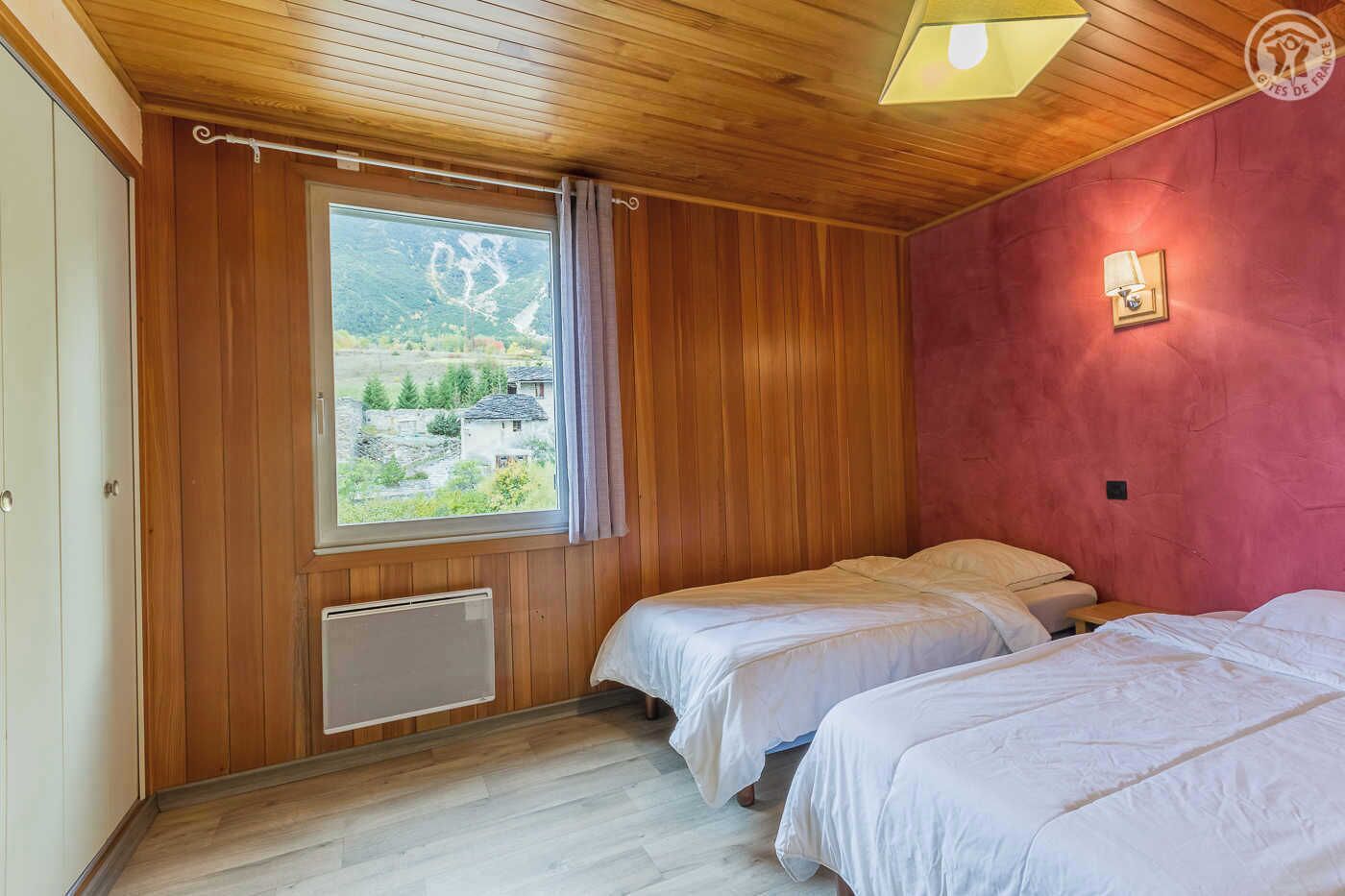 Une chambre avec deux lits, des murs en bois, une fenêtre donnant sur un paysage verdoyant et des bâtiments, et un luminaire au plafond.