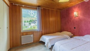 Une chambre avec deux lits, des murs en bois, une fenêtre donnant sur un paysage verdoyant et des bâtiments, et un luminaire au plafond.