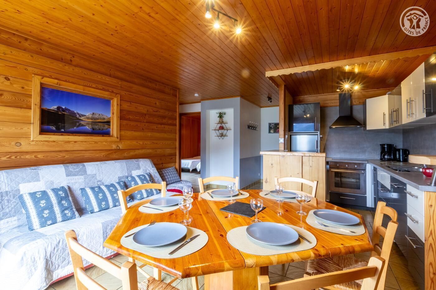 L’intérieur chaleureux d’un chalet avec une table à manger en bois, préparée pour quatre personnes, un canapé agrémenté de coussins à motifs et un coin cuisine équipé d’appareils électroménagers modernes.