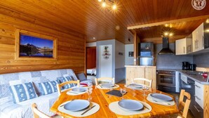 L’intérieur chaleureux d’un chalet avec une table à manger en bois, préparée pour quatre personnes, un canapé agrémenté de coussins à motifs et un coin cuisine équipé d’appareils électroménagers modernes.
