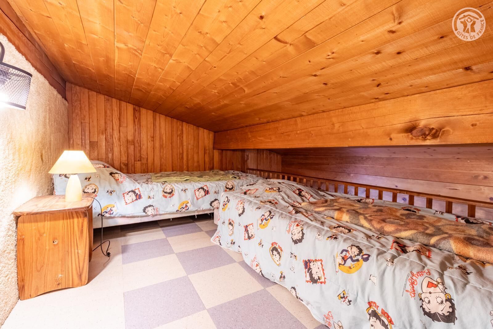 A wooden room with a checkered floor, a bed with cartoon character bedding, a nightstand with a lamp, and a window with a mesh screen.