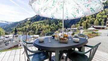 Een ronde tafel met stoelen, een parasol en eten op een terras met uitzicht op een berg.