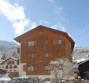 Un bâtiment en bois à plusieurs étages, doté d’un toit en pente, vraisemblablement un hôtel ou un chalet, entouré d’arbres et de montagnes enneigées en arrière-plan.