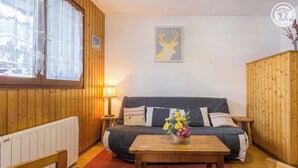 Een gezellige kamer met een houten paneelwand, een grijze bank met kussens, een houten salontafel met een bloemenvaas, en een afbeelding van een hertenhoofd in een lijst aan de wand.
