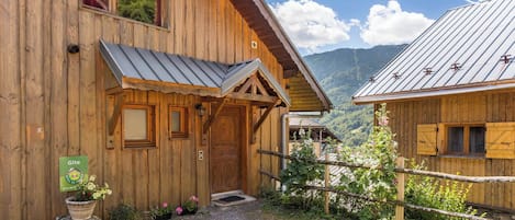 Een houten chalet met een metalen dak, een houten deur en een klein terras. Er hangt een bordje bij de deur en er staat een bloempot op de grond.